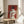 May include: Framed poster print on wooden side table next to beige upholstered chair and plant. Artwork shows woman from behind with dark hair in bun, wearing white draped clothing, holding red roses against burgundy background. Natural lighting creates soft shadows across cream wall.
