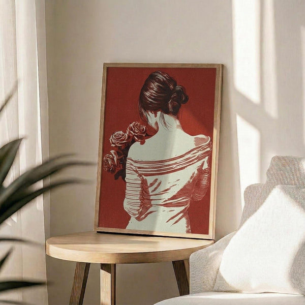 May include: Framed poster print on wooden side table next to beige upholstered chair and plant. Artwork shows woman from behind with dark hair in bun, wearing white draped clothing, holding red roses against burgundy background. Natural lighting creates soft shadows across cream wall.