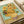 May include: Van Gogh's famous Sunflowers painting reproduction displayed in an open art book or portfolio, showing vibrant yellow sunflowers in a ceramic vase against a turquoise background. The print sits on white pages with cream ribbon visible, photographed on a light wooden surface with soft natural lighting highlighting the rich brushstroke textures and warm golden tones.