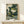 May include: Framed poster print displayed on light wood console table against white wall. Artwork shows anthropomorphic green frog wearing dark robes, smoking wooden pipe with white smoke swirls, surrounded by stylized ocean waves in traditional Japanese ukiyo-e woodblock print style. Teal and cream color palette with intricate wave patterns. Small wrapped gift box sits beside frame.