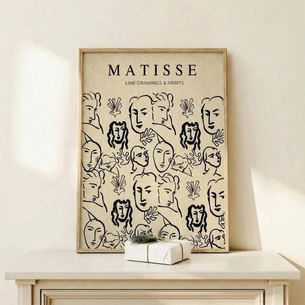 May include: Framed beige poster with black line drawings in Matisse's distinctive style, featuring repeated faces and botanical elements in a pattern. Light wood frame on white wooden shelf against cream wall, with small wrapped gift box beside it.