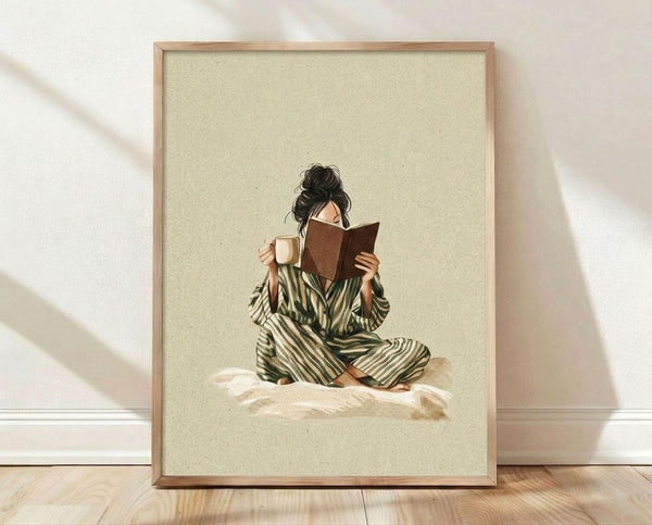 May include: Framed art print in natural wood frame leaning against white wall on light wooden floor with soft shadow patterns. The artwork shows a woman with dark hair in a messy bun, wearing green and cream striped pajamas or robe, sitting cross-legged on rumpled white bedding while reading a brown book and holding a cream-colored mug. The illustration has a warm, cozy aesthetic with muted earth tones on a sage green textured background.