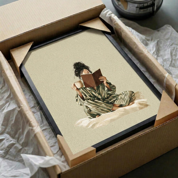 May include: Framed art print showing woman in green striped robe reading book while sitting cross-legged on neutral beige background, displayed in black frame being unpacked from cardboard shipping box with protective wrapping and packing materials on white surface