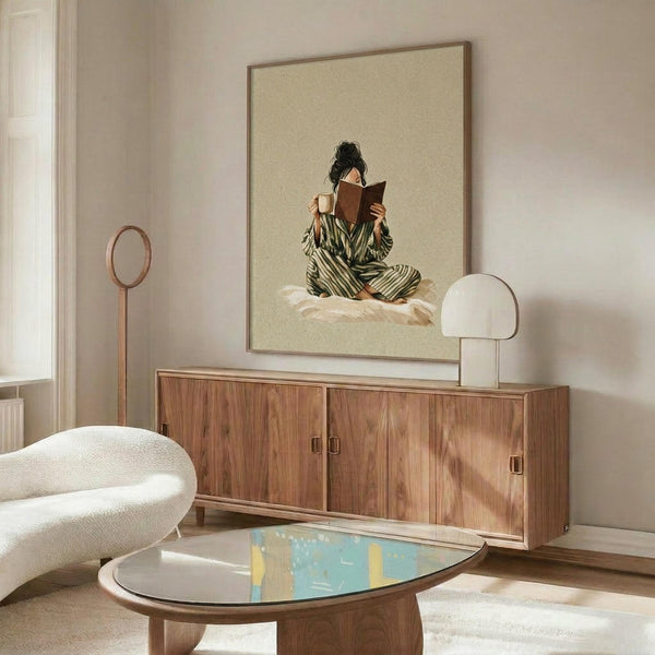 May include: Framed artwork showing a woman in green striped pajamas reading a book while holding a mug, sitting cross-legged on white bedding against a beige background. The print hangs above a wooden mid-century modern credenza with sliding doors in a minimalist living room with white walls, cream chair, oval glass coffee table, and modern lamp.