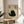 May include: Framed artwork featuring a black cat illustration with yellow eyes on beige textured background, accompanied by Japanese characters and red seal stamp, displayed on light wood round side table next to beige upholstered chair with white pillow, plant leaves visible in corner, natural lighting creating soft shadows on neutral wall