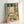 May include: Framed reproduction of Matisse's Fauvist painting showing an open window view of a harbor with sailing boats, rendered in vibrant greens, pinks, oranges and blues. The artwork features bold brushstrokes depicting window frames, potted plants on the sill, and a Mediterranean coastal scene beyond. The print is displayed in a light wooden frame, leaning against a white wall corner.