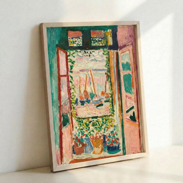 May include: Framed reproduction of Matisse's Fauvist painting showing an open window view of a harbor with sailing boats, rendered in vibrant greens, pinks, oranges and blues. The artwork features bold brushstrokes depicting window frames, potted plants on the sill, and a Mediterranean coastal scene beyond. The print is displayed in a light wooden frame, leaning against a white wall corner.