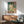 May include: Colorful Matisse fauve painting print showing open window with view of boats on water, displayed in thin gold frame above wooden sideboard. Mid-century modern room with sage green upholstered chair, white ceramic table lamp, and round wood coffee table. Bright sunlight streams across white walls creating dramatic shadows.
