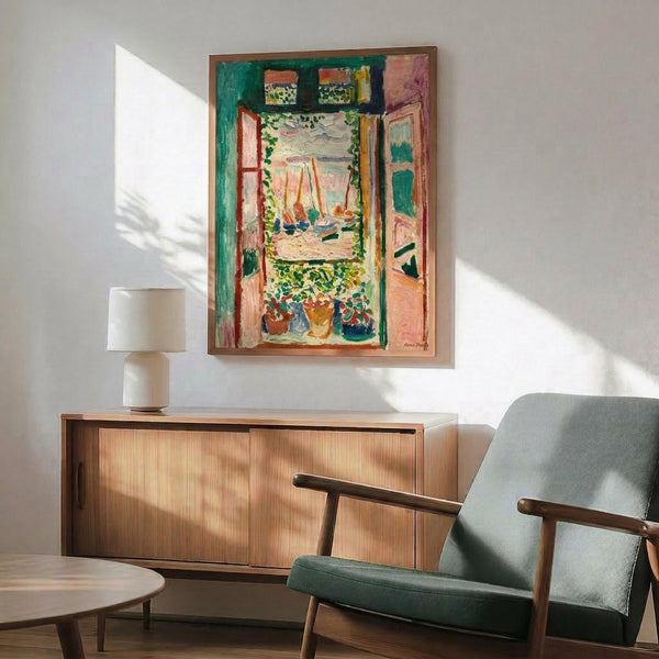May include: Colorful Matisse fauve painting print showing open window with view of boats on water, displayed in thin gold frame above wooden sideboard. Mid-century modern room with sage green upholstered chair, white ceramic table lamp, and round wood coffee table. Bright sunlight streams across white walls creating dramatic shadows.