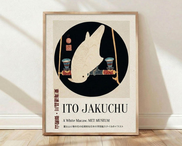 May include: Framed poster print leaning against white wall on wooden floor. Features circular black background with white macaw illustration in traditional Japanese art style, flanked by decorative vessels. Text reads "ITO JAKUCHU" and "A White Macaw. MET MUSEUM" with Japanese characters. Natural wood frame with white matting, soft window light creating shadows on textured white wall.