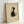 May include: Framed artwork displaying a minimalist black and white cat illustration in Japanese ink wash style, sitting and looking upward with whiskers detailed. The cat has dark fur on back and white chest. Red artist seal stamp in bottom right corner. Cream textured background in light wood frame, leaning against white wall.