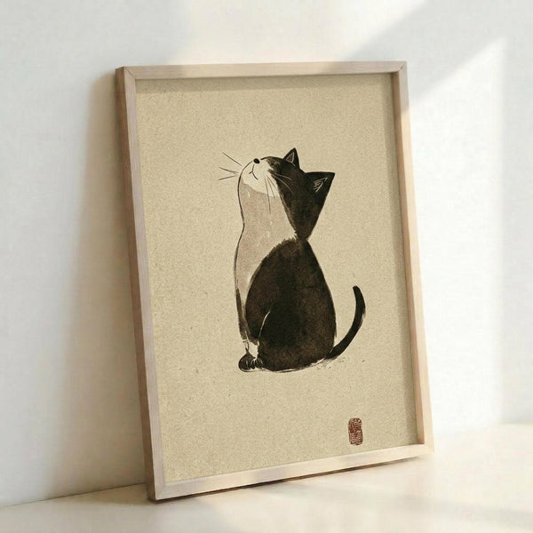 May include: Framed artwork displaying a minimalist black and white cat illustration in Japanese ink wash style, sitting and looking upward with whiskers detailed. The cat has dark fur on back and white chest. Red artist seal stamp in bottom right corner. Cream textured background in light wood frame, leaning against white wall.
