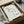 May include: Minimalist line drawing of a flamingo signed "Picasso" displayed in a black frame within an unboxing scene. The artwork features simple black curved lines forming a stylized flamingo on cream paper. The framed piece sits in brown cardboard packaging with white tissue paper, surrounded by wooden protective corners. A glass vessel is partially visible in the background on a neutral surface.