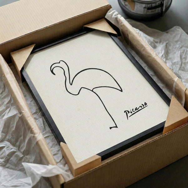 May include: Minimalist line drawing of a flamingo signed "Picasso" displayed in a black frame within an unboxing scene. The artwork features simple black curved lines forming a stylized flamingo on cream paper. The framed piece sits in brown cardboard packaging with white tissue paper, surrounded by wooden protective corners. A glass vessel is partially visible in the background on a neutral surface.