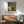 May include: Framed botanical poster featuring a colorful hummingbird with spread wings hovering near large pink and cream flowers surrounded by lush green foliage. The vintage-style artwork has rich yellows, greens, and coral tones in an ornate Victorian botanical illustration style. Displayed above a wooden sideboard with sliding doors next to a sage green mid-century armchair with wooden arms in a modern living room with white walls and natural lighting.