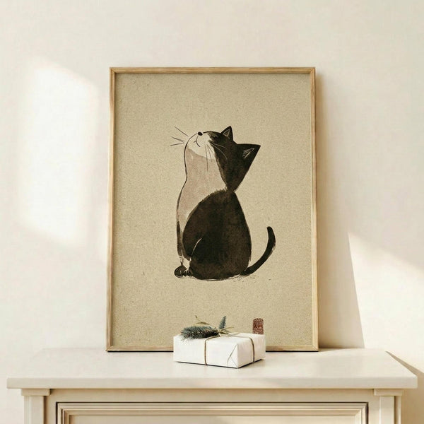 May include: Framed minimalist cat artwork in light wood frame displayed on pale wooden surface against cream wall. Black and white tuxedo cat sits looking upward in Japanese ink wash style on beige textured background. Small wrapped white gift box with evergreen sprig sits beside frame. Soft natural lighting creates gentle shadows.