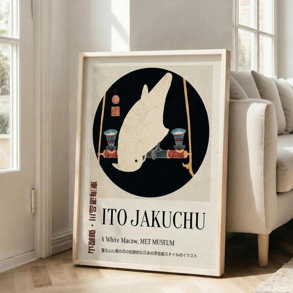 May include: Framed poster featuring a white macaw in profile within a black circle on cream background, displayed in bright living room corner next to beige sofa. The artwork shows Japanese-style illustration with decorative elements and text reading "ITO JAKUCHU" and "A White Macaw. MET MUSEUM" below. Natural wood frame leans against white wall near tall windows with sunlight streaming across hardwood floors.
