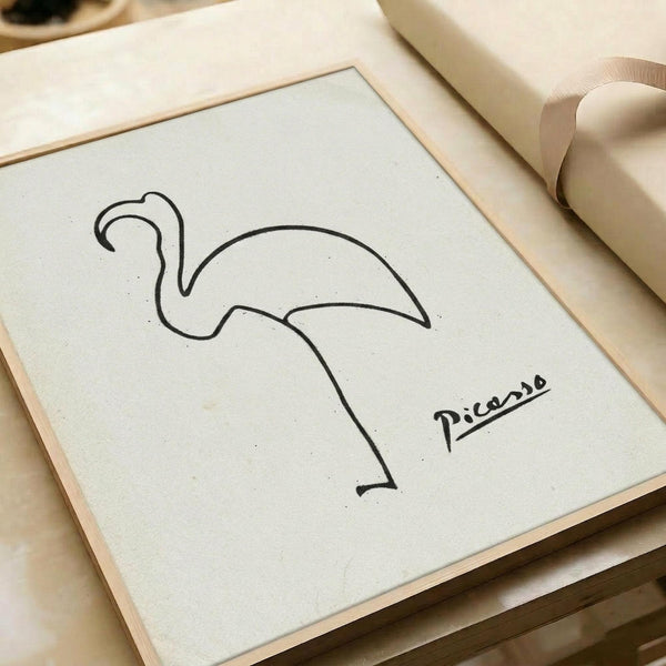 A minimalist line drawing of a flamingo created with simple black curved lines on white paper, signed "Picasso" in the bottom right corner. The artwork is displayed in a light wood frame and sits on a neutral beige surface alongside rolled paper or fabric materials, creating a clean, artistic workspace aesthetic.