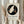 May include: Person in beige ruffled sleeves holding framed poster showing white macaw in black circle with decorative vases. Light wood frame with cream matting, dried pampas grass visible to left, soft natural lighting against neutral curtained background.