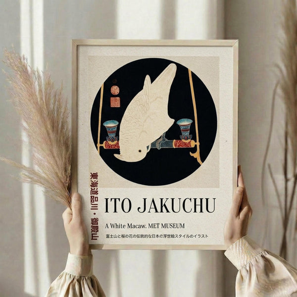 May include: Person in beige ruffled sleeves holding framed poster showing white macaw in black circle with decorative vases. Light wood frame with cream matting, dried pampas grass visible to left, soft natural lighting against neutral curtained background.