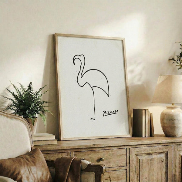 May include: Minimalist line drawing of a flamingo in black ink on white paper, signed "Picasso" in cursive, displayed in a light wood frame on a rustic wooden sideboard. The simple, continuous-line artwork shows the bird's curved neck and body. Scene includes a cream upholstered chair, fern plant, stacked books, and ceramic vase in warm, neutral-toned living room setting.