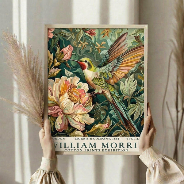 May include: Hands holding a framed vintage-style botanical art print featuring a colorful hummingbird with spread wings hovering near large peach and pink flowers against a dark green leafy background. The print has "William Morris Cotton Prints Exhibition" text at bottom and sits in a light wooden frame. Dried pampas grass visible to the left against cream curtains in soft natural lighting.