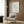 May include: Minimalist line drawing of a flamingo signed Picasso displayed in light wood frame on warm wood paneled wall above cream upholstered armchair with geometric patterned pillow, brass floor lamp, and dark round side table with open book in modern neutral-toned living room setting