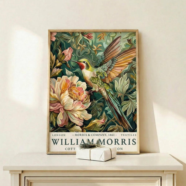 May include: Framed vintage-style botanical poster featuring a colorful hummingbird with red throat and yellow-green plumage hovering near large pink peony flowers surrounded by lush green foliage. The artwork has rich, detailed illustration style with text at bottom reading "London Morris & Company 1861 Textiles William Morris Collection." Natural wood frame sits on light wooden shelf next to small white wrapped gift box against cream colored wall.
