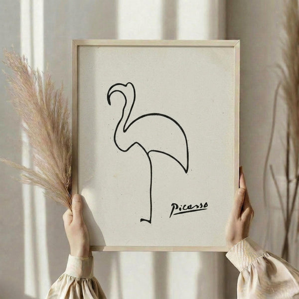 May include: Person in beige ruffled sleeve top holding framed minimalist line drawing of a flamingo in simple black strokes against cream background, with dried pampas grass stems visible beside the light wood frame, photographed in soft natural lighting near white curtained window