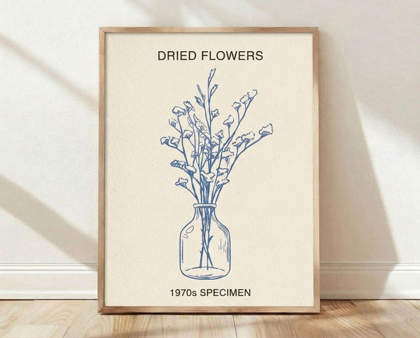 May include: Framed botanical poster print leaning against white wall on wooden floor. Light wood frame contains cream-colored artwork with blue line drawing of dried flowers in glass vase. Text reads "DRIED FLOWERS" at top and "1970s SPECIMEN" at bottom. Natural lighting creates soft shadows on textured white wall background.