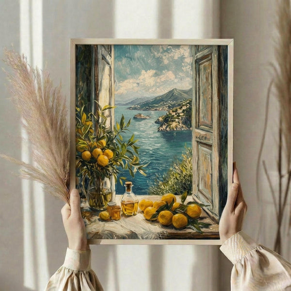 May include: Person holding framed art print showing Mediterranean coastal scene through open window. Painting features turquoise waters, hillside villages, lemon tree with bright yellow fruit, glass bottles, and scattered lemons on white windowsill. Warm beige wooden frame against neutral wall with dried pampas grass visible on left side.