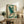 May include: Framed Japanese-style artwork featuring a blue-green heron with outstretched wings flying through stylized cream and teal clouds, displayed on a rustic wooden console table alongside books, a ceramic vase, and a fern plant in a modern living room with neutral walls and leather seating.