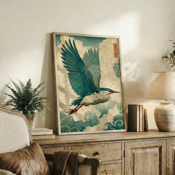 May include: Framed Japanese-style artwork featuring a blue-green heron with outstretched wings flying through stylized cream and teal clouds, displayed on a rustic wooden console table alongside books, a ceramic vase, and a fern plant in a modern living room with neutral walls and leather seating.