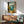 May include: Framed Mediterranean coastal artwork showing open window view of turquoise sea, mountains, and citrus fruits on windowsill, displayed above wooden sideboard with white lamp in modern living room with mint green armchair and natural lighting creating geometric shadows on white wall.