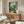 May include: Mediterranean coastal scene painting displayed above wooden sideboard in modern living room. Artwork shows open window view of turquoise water, mountains, and lemon tree with oranges on windowsill. Room features cream walls, white curved chair, glass coffee table, and brass table lamps. Painting has vintage European style with blue skies and rustic shutters.
