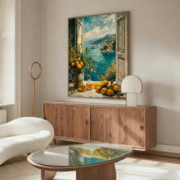 May include: Mediterranean coastal scene painting displayed above wooden sideboard in modern living room. Artwork shows open window view of turquoise water, mountains, and lemon tree with oranges on windowsill. Room features cream walls, white curved chair, glass coffee table, and brass table lamps. Painting has vintage European style with blue skies and rustic shutters.