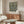 May include: William Morris botanical poster print in thin gold frame hanging above mid-century walnut credenza. Artwork features intricate black, white and olive green floral pattern with large blooms and detailed foliage. Modern living room setting with cream curved chair, glass-top coffee table, and minimalist brass floor lamp on neutral walls.