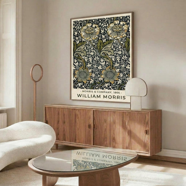 May include: William Morris botanical poster print in thin gold frame hanging above mid-century walnut credenza. Artwork features intricate black, white and olive green floral pattern with large blooms and detailed foliage. Modern living room setting with cream curved chair, glass-top coffee table, and minimalist brass floor lamp on neutral walls.