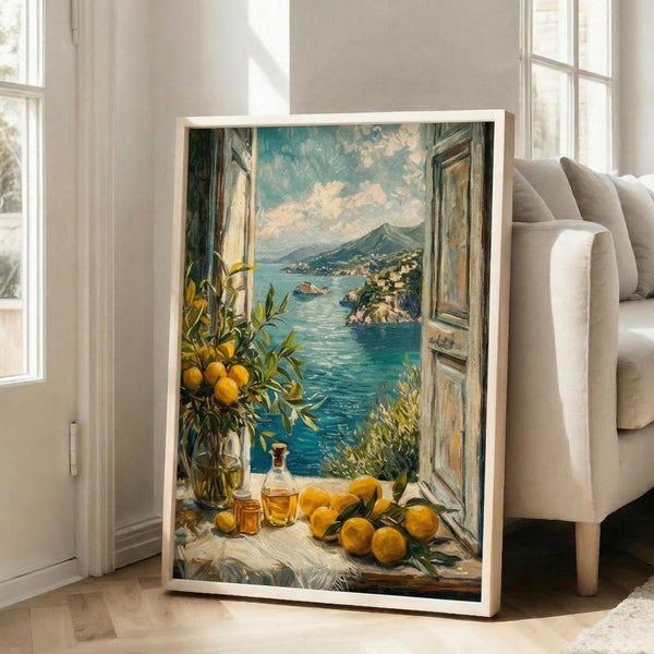 May include: Framed poster print leaning against white wall next to beige sofa in bright living room. Artwork shows Mediterranean coastal scene through open wooden window shutters with turquoise blue water, mountain, and hillside village. Foreground features yellow lemons, citrus branches in vase, and glass bottles on white windowsill. Painted in impressionistic style with warm yellows and cool blues.
