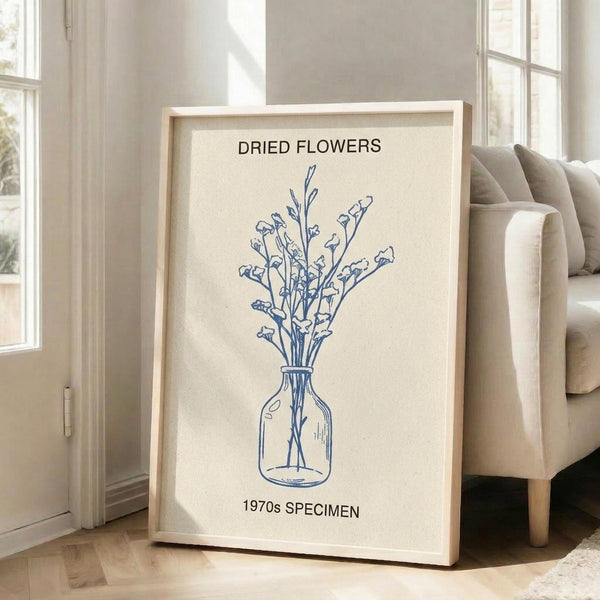 May include: Framed botanical wall art with cream background and navy blue line drawing of dried flowers in a glass vase, displayed on light wooden floors next to a beige upholstered chair in a bright room with white walls and large windows