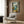 May include: Mediterranean coastal artwork displayed in modern living room with beige armchair, brass floor lamp, and wood paneled wall. The framed print shows an open window view of turquoise waters, mountains, and lemon branches with oranges and bottles on the windowsill. Warm natural lighting creates cozy reading nook atmosphere.