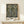 May include: Framed vintage floral pattern poster with intricate black background featuring ornate cream and olive green flowers and leaves in William Morris decorative style, displayed on light wood shelf with small wrapped gift box, against neutral beige wall in minimalist interior setting.
