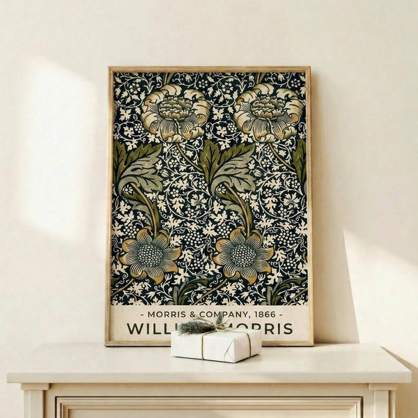 May include: Framed vintage floral pattern poster with intricate black background featuring ornate cream and olive green flowers and leaves in William Morris decorative style, displayed on light wood shelf with small wrapped gift box, against neutral beige wall in minimalist interior setting.