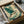 May include: Framed Japanese-style artwork of a blue-green heron with outstretched wings flying through stylized cream and teal clouds and waves, displayed in a black frame being carefully unpacked from a cardboard shipping box with protective tissue paper wrapping.