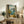 May include: Framed art print leaning against white wall on rustic wooden sideboard, showing Mediterranean coastal scene viewed through open window with yellow lemons, blue water, and mountains. Room styled with fern plant, books, ceramic vases, and cream upholstered furniture in warm neutral tones.