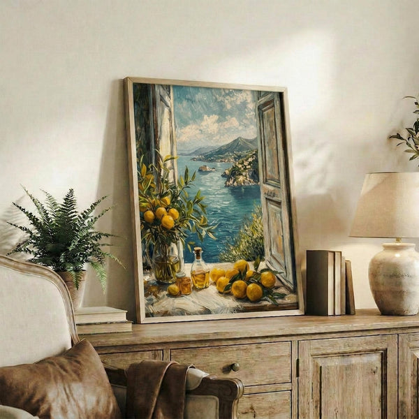 May include: Framed art print leaning against white wall on rustic wooden sideboard, showing Mediterranean coastal scene viewed through open window with yellow lemons, blue water, and mountains. Room styled with fern plant, books, ceramic vases, and cream upholstered furniture in warm neutral tones.