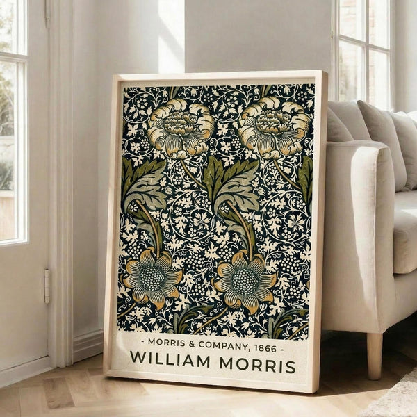 May include: Framed poster with William Morris floral pattern in dark navy background featuring ornate golden flowers, green leaves, and intricate white botanical details. Light wood frame leans against white wall next to beige upholstered chair in bright room with large windows and natural lighting on hardwood floors.