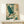 May include: Japanese-style artwork showing a blue-green heron with outstretched wings flying through stylized cream-colored clouds and teal waves. The print features traditional woodblock print aesthetics with detailed feather textures and flowing water patterns. Displayed in a thin wooden frame on a light wood shelf next to a small white wrapped gift box against a cream wall.