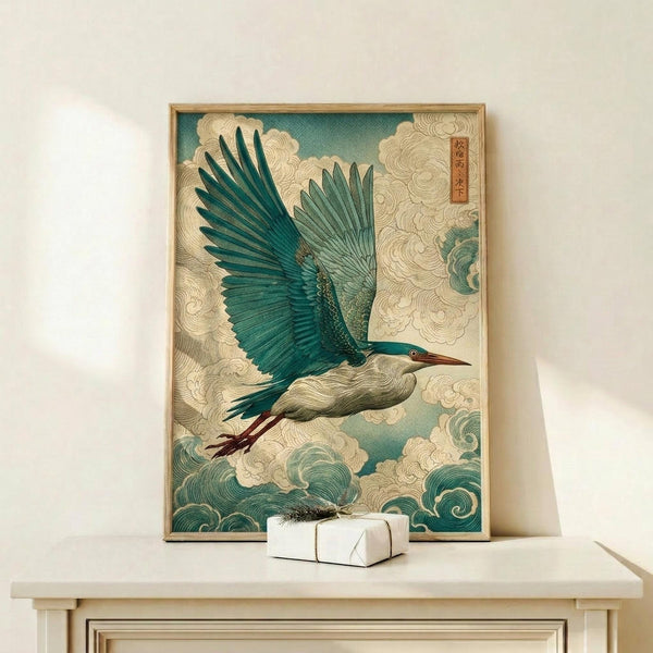May include: Japanese-style artwork showing a blue-green heron with outstretched wings flying through stylized cream-colored clouds and teal waves. The print features traditional woodblock print aesthetics with detailed feather textures and flowing water patterns. Displayed in a thin wooden frame on a light wood shelf next to a small white wrapped gift box against a cream wall.