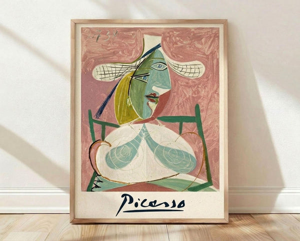 May include: A framed Picasso-style cubist portrait showing a seated woman with geometric features in green, yellow and pink tones against a coral background, displayed in a light wood frame on wooden flooring against white walls with natural lighting creating soft shadows.